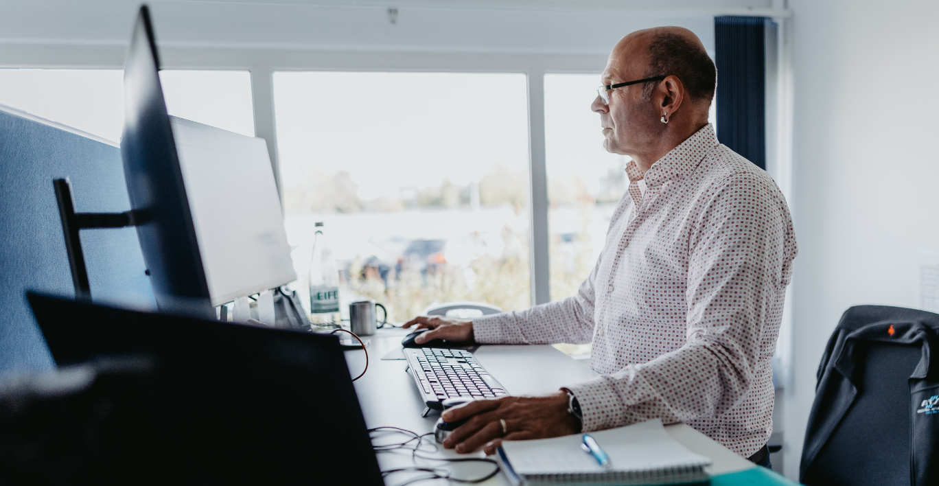 Man in a white shirt working on a computer in an open office with a window in the background