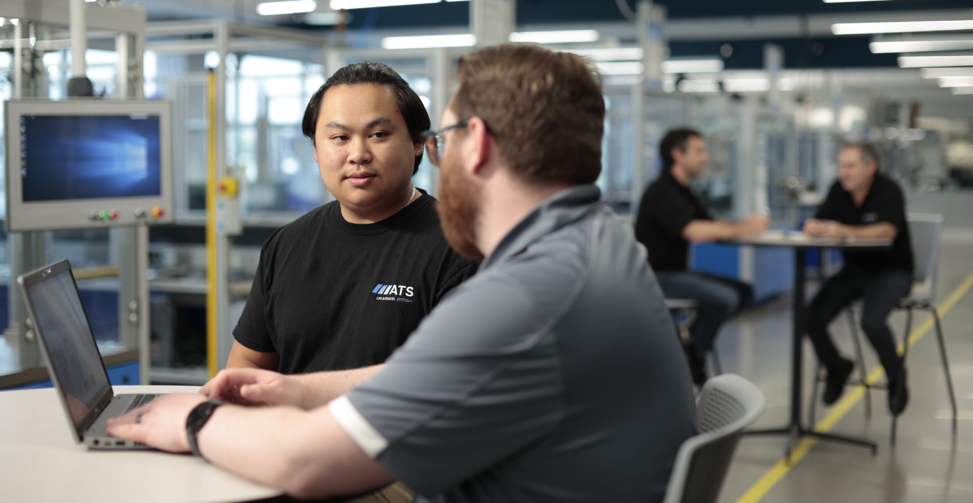 Two men sitting at a table near a manufacturing environment talking