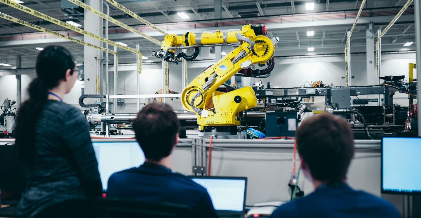Three individuals looking at a large yellow robot with three computer screens shown, meant to showcase a remote manufacturing environment. 